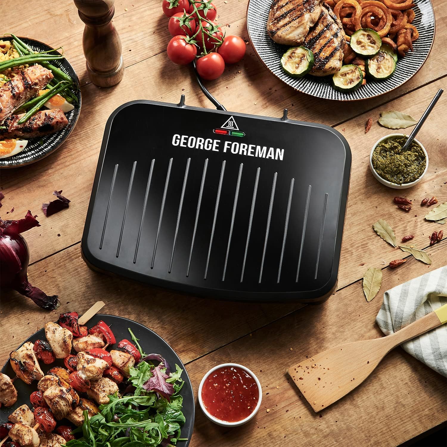 A George Foreman electric grill on a wooden table surrounded by grilled vegetables, garnishes, and sauces, showcasing a variety of healthy meals.