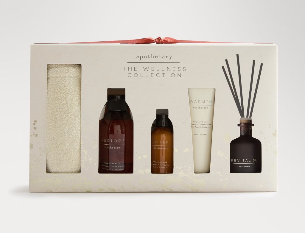 Wellness gift set featuring four products: 'Restore' body wash, 'Sleep' body oil, 'Warmth' hand cream, and 'Revitalise' diffuser, along with a white towel, packaged in a decorative box.